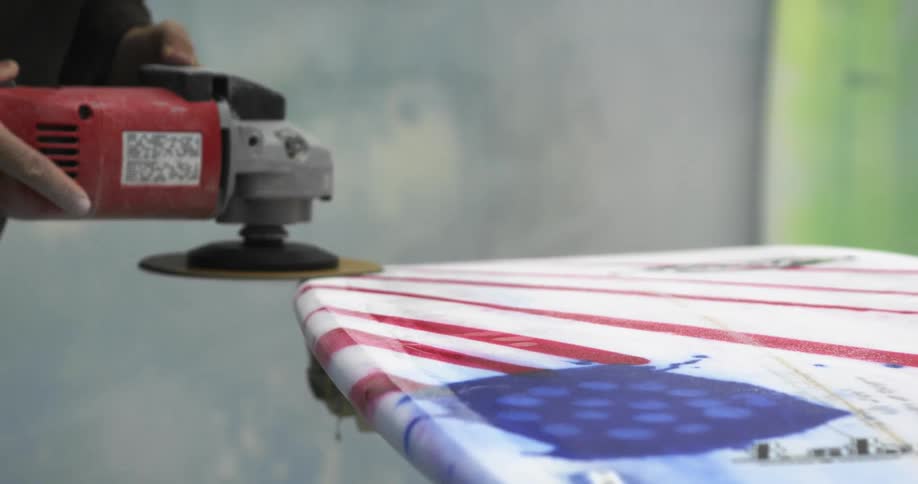 A man uses a sander to smooth a surfboard painted with an american flag design