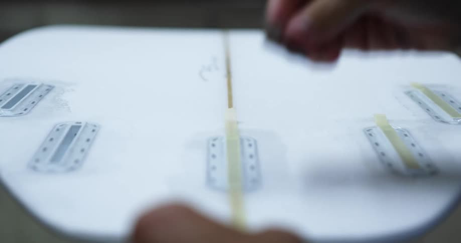 A person crafting a surfboard places tape on the surface and cuts it with a razor