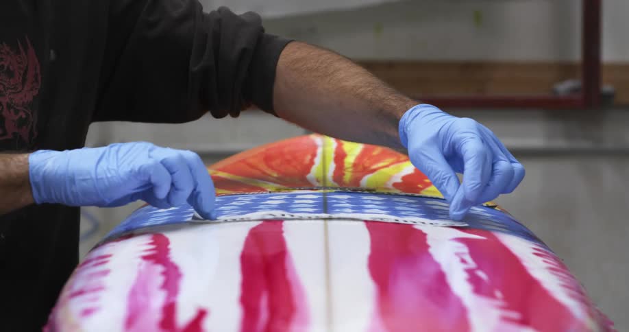 A man wearing gloves taps a strip into place on a freshly painted surfboard