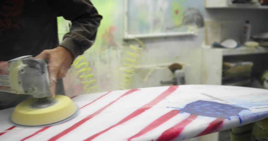 A man moves a spinning electric polishing tool back and forth smoothing out the surface of a surfboard on a table in a workshop