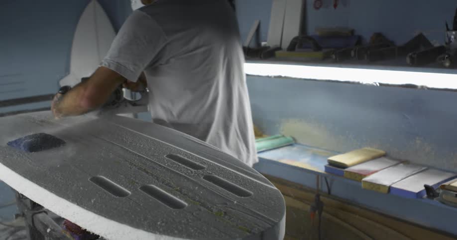 A man wearing a respirator mask uses a power tool to smooth and shape a skim board