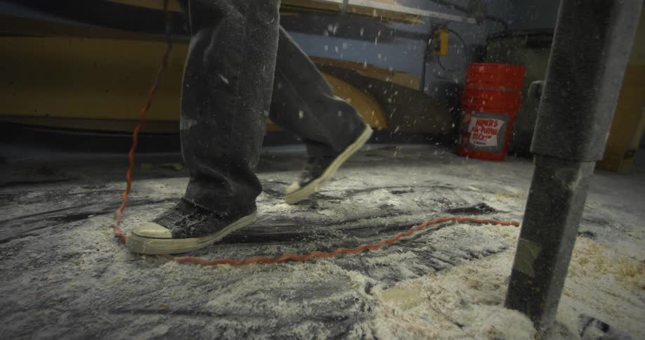 Sneakered feet walk through sawdust on the floor as more sawdust falls from the use of a power sander