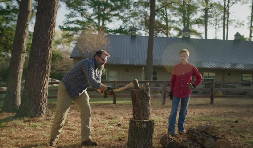 Father with glasses a beard a blue shirt and beige pants shows his son wearing a red shirt and jeans how to cut wood with an ax