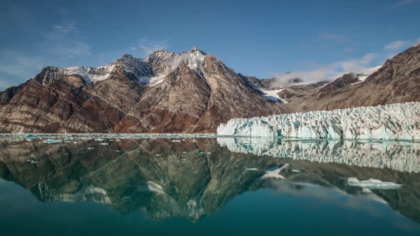 A lake with water and ice with a mountain range the sky and clouds in the background
