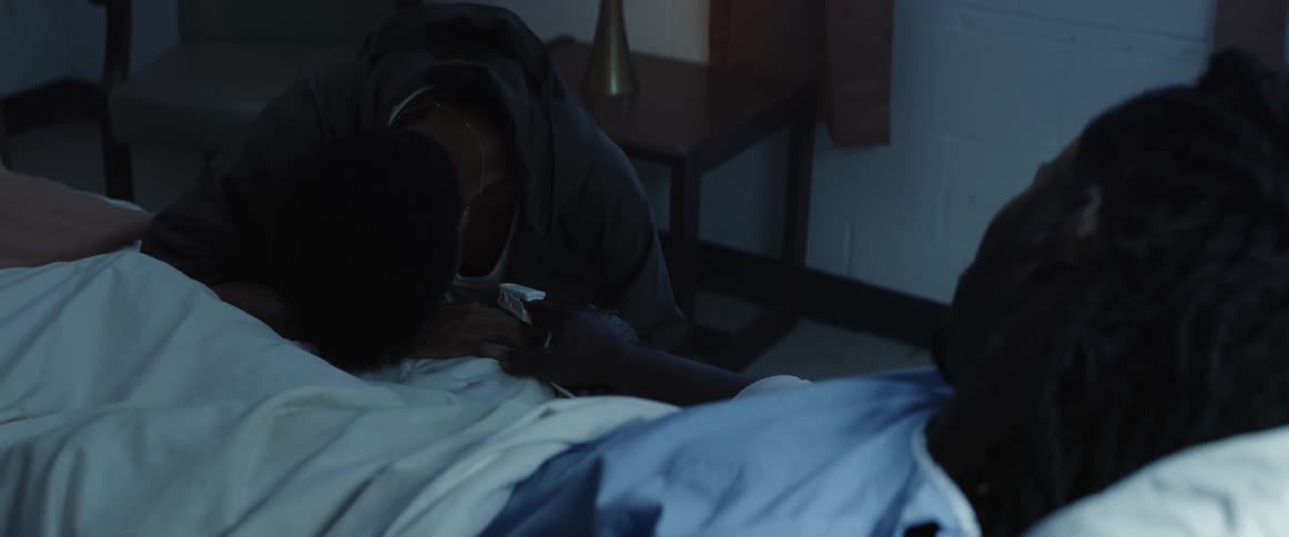 A woman kneels at a hospital bed and cries as the patient reaches his hand out and holds her hand