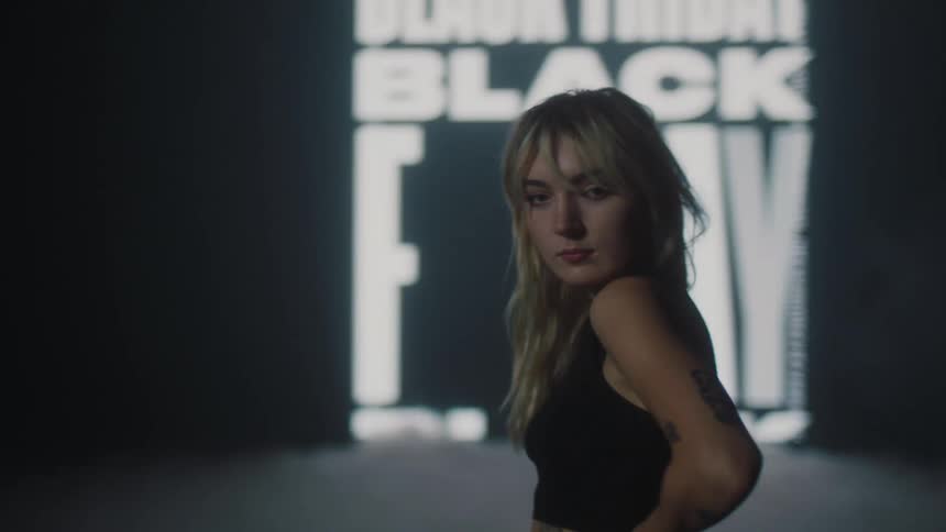 Blonde woman moves around and poses on fog covered stage backed by large display