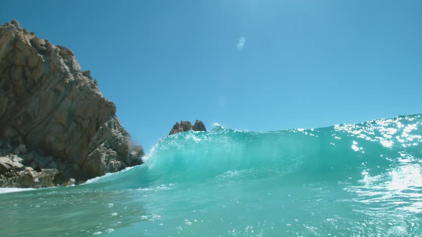 A large turquoise ocean wave curls near a rock formation on the shore on a sunny day