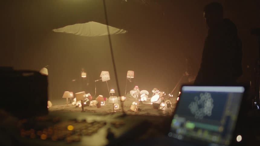 A man stands by watching as men sweep and spread sand around table and floor lamps for a video shoot