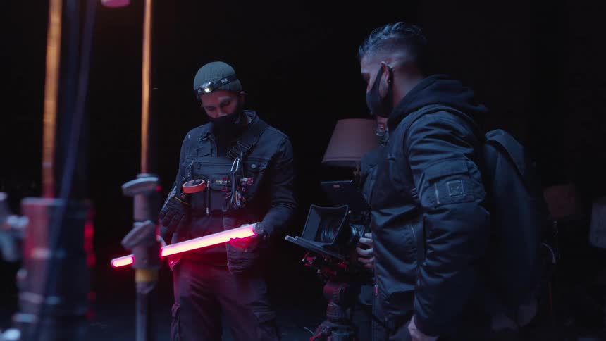 Two men stand at the sides of another man who is in the middle holding a professional grade video camera while one of them holds a red florescent light bulb stick