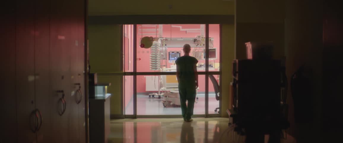 A nurse walking on a dark hallway in a hospital stops in front of a patients room