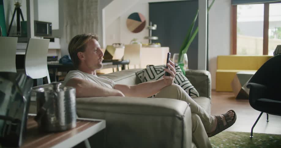 A man sitting in the couch on the living room greeting someone on a video call in his smartphone