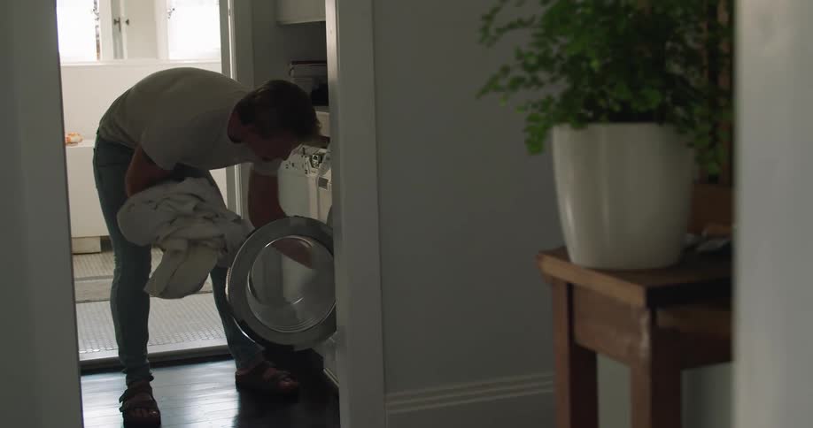 A man in a hallway putting laundry in a washing machine with a doorway in the background
