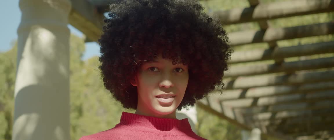 A woman with big natural curls poses outside under a pergola on a sunny day