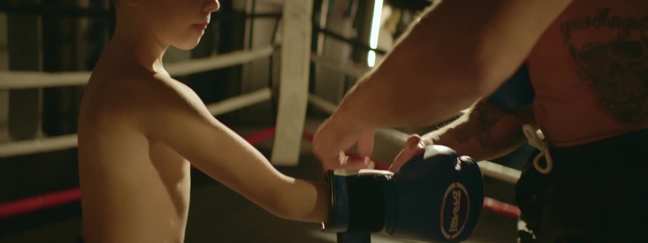 A strong shirtless muscular man helps a boy putting boxing gloves inside a ring