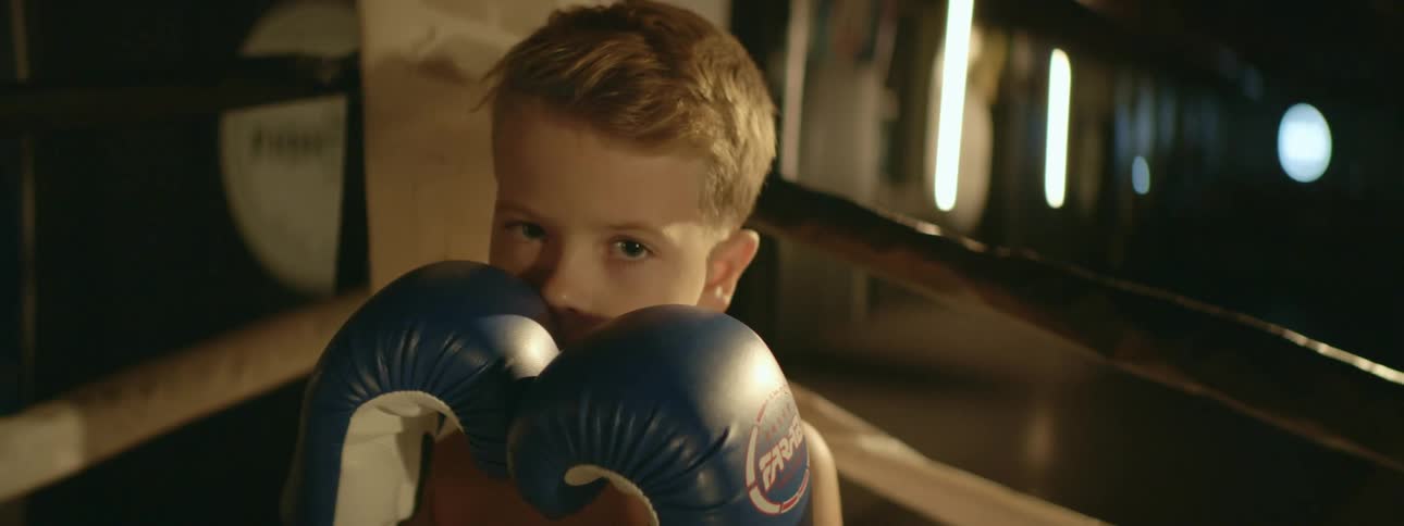 Blonde boy standing in boxing ring holds glove covered hands near mouth