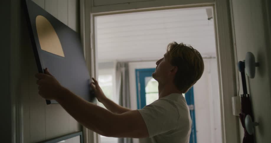 A man walks down a narrow hallway in his house and hangs an art piece on the wall