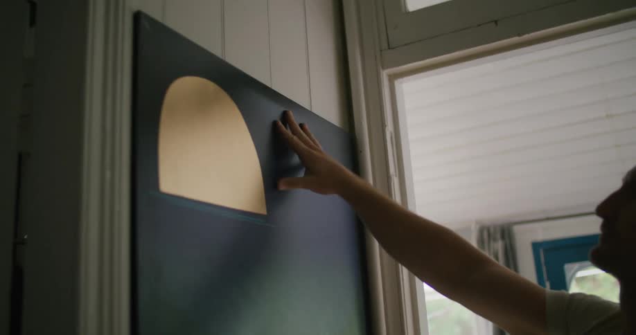 A man at home hangs art work on a wall and looks at it