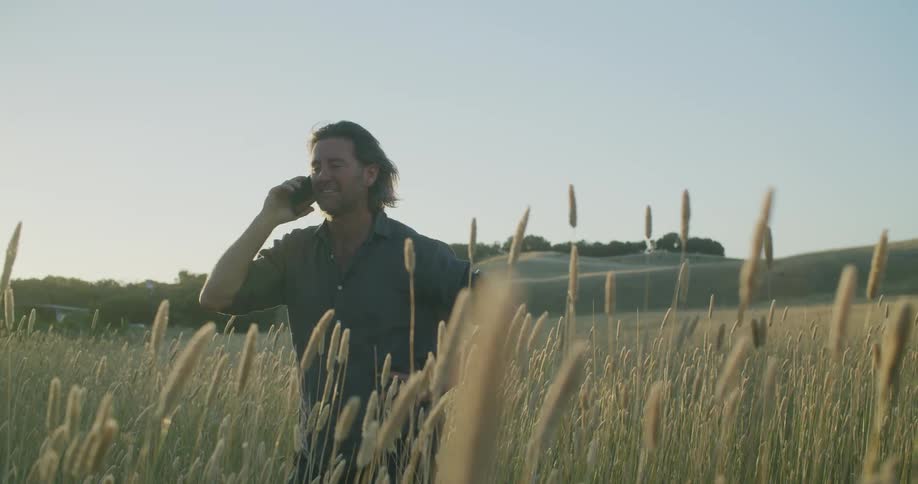 A man laughs on the phone while standing in a meadow surrounded with tall willow reeds