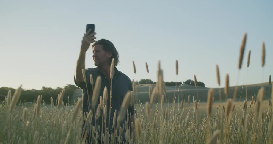 A man standing on a field next to the hills trying to get signal for his phone at sunset