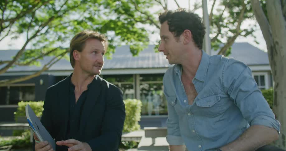 Two men sit outside a house and talk and look at a laptop