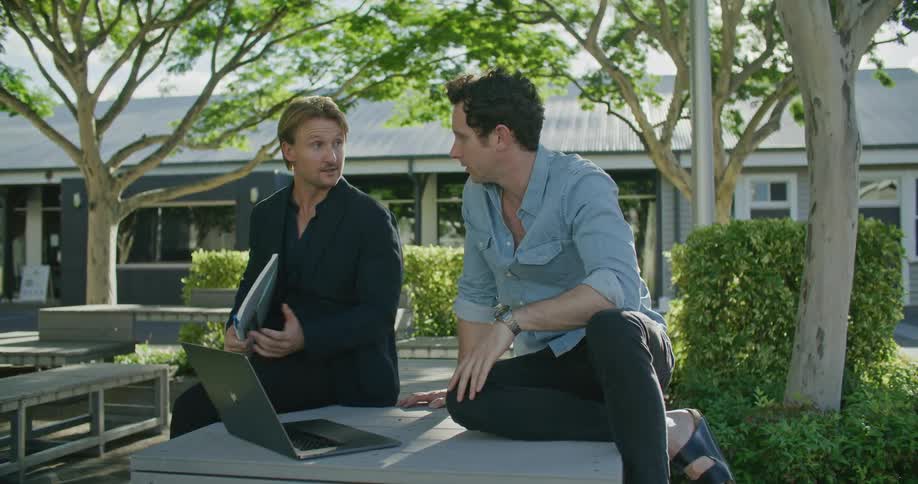 A pair of men talk together while looking at a laptop as they sit outdoors during the day