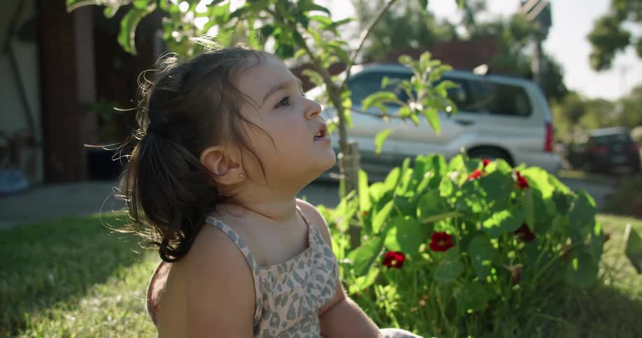 A baby girl sitting on grass with plants trees the sky and clouds in the background