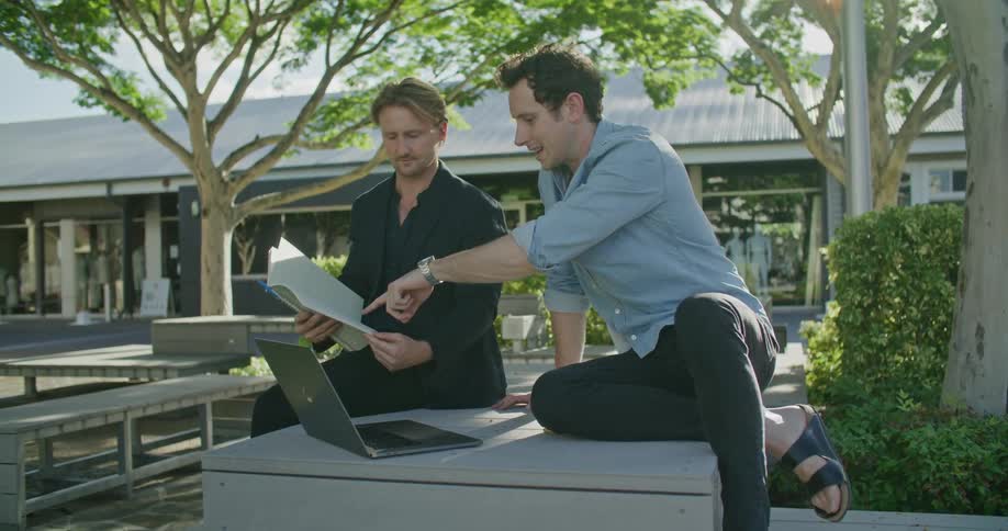 Two men talking outside while referencing a notebook and laptop