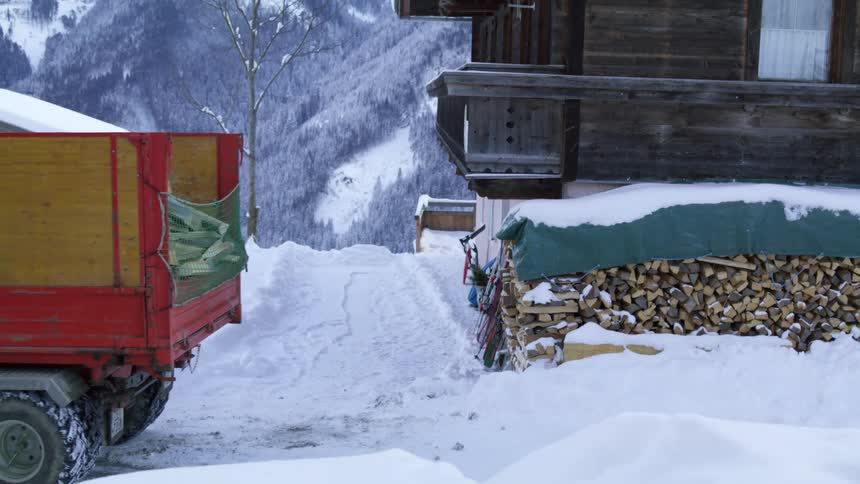 A truck carrying chopped lumber backs toward a stack of firewood covered by a tarp on a snowy day