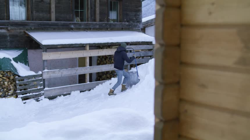 A man pushes a wheelbarrow style shovel outside his cabin and dumps snow onto a pile
