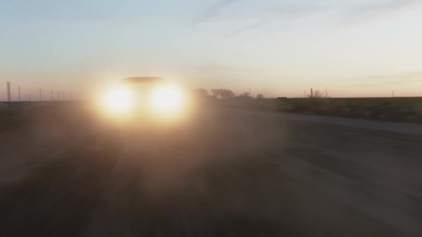 A vehicle kicks up dust as it speeds down a rural dirt road at dusk