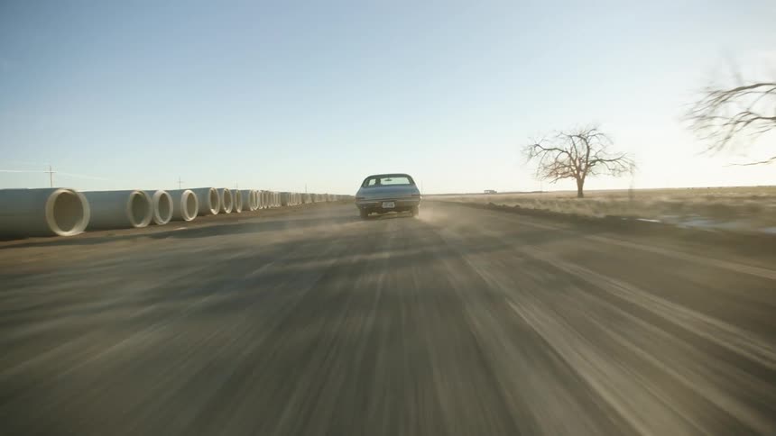 A car driving on a road raising dust with the sky and clouds in the background