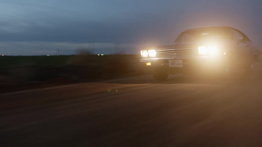 A blue vintage muscle car with headlights on driving on a dusty rural road at sunset