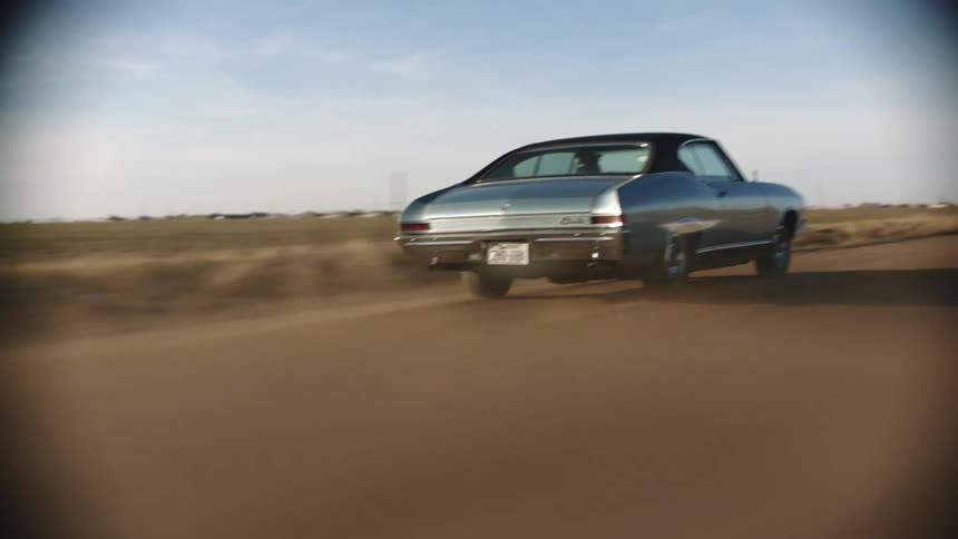 A car driving on a road next to a grassy field with the sky and clouds in the background