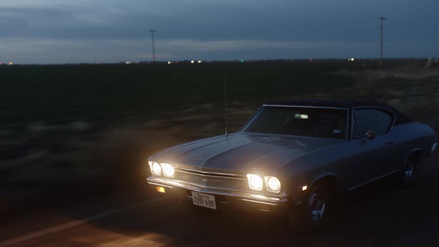 An antique car traveling on a rural road at night with illuminated headlights