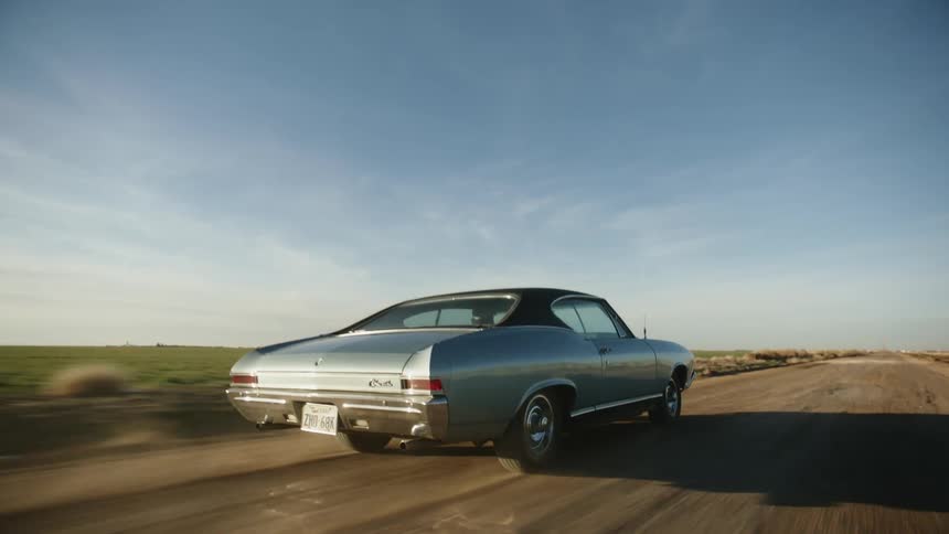 A blue vintage muscle car driving on a dusty rural road at sunset