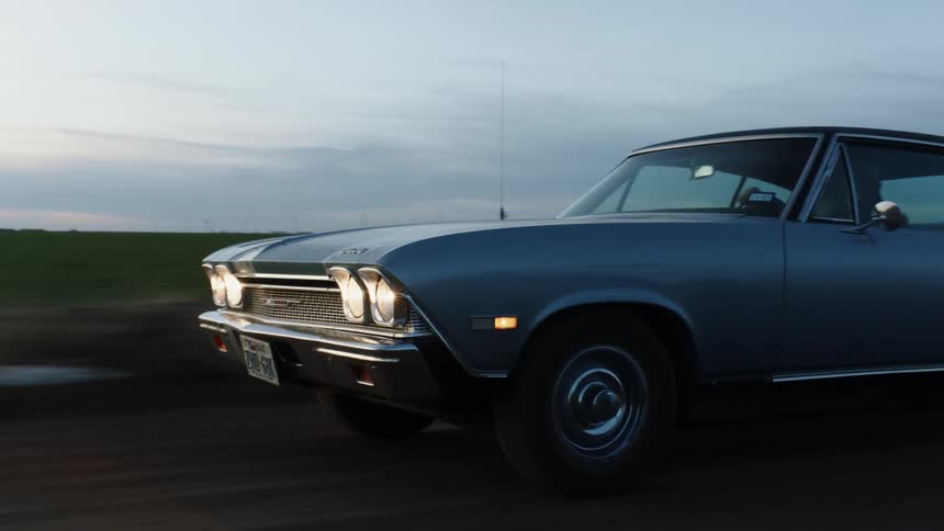 A man driving a car on a road next to a grassy field with the sky and clouds in the background