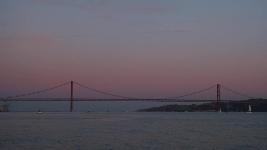 A suspension bridge over the boats sailing on a river at sunset