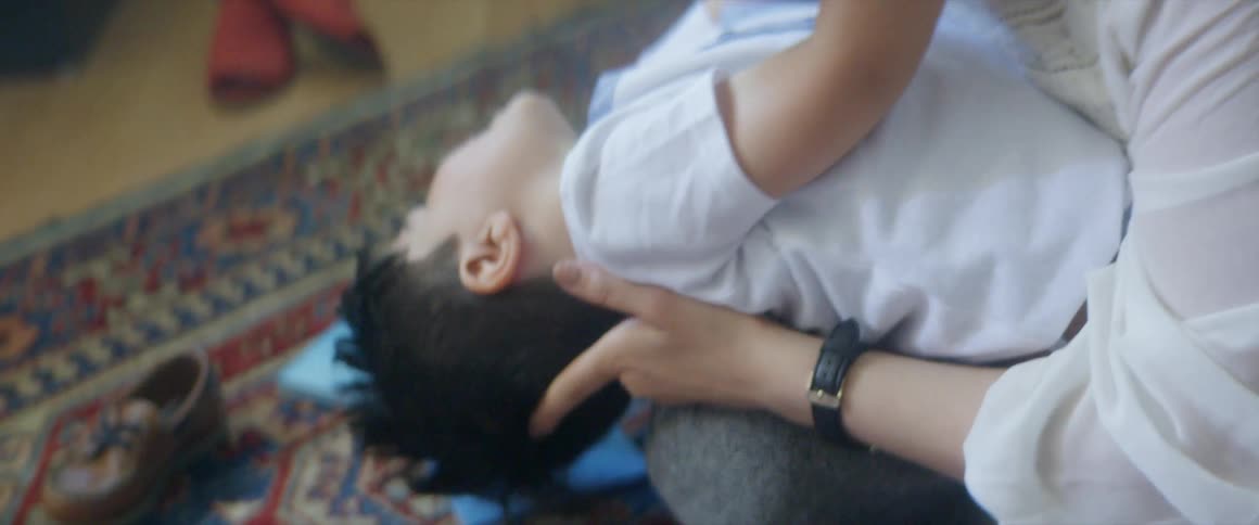 Mother holding son in her lap tickles him until he falls back then picks him up in living room with plants and books behind them and books and shoes on the rug and floor beside them