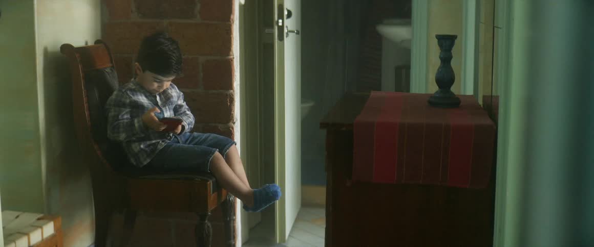 A little boy sits on a chair in a room next to an opened door looking at a phone held in his hands