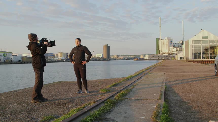 A camera man holds a camera while a woman standing near a river speaks