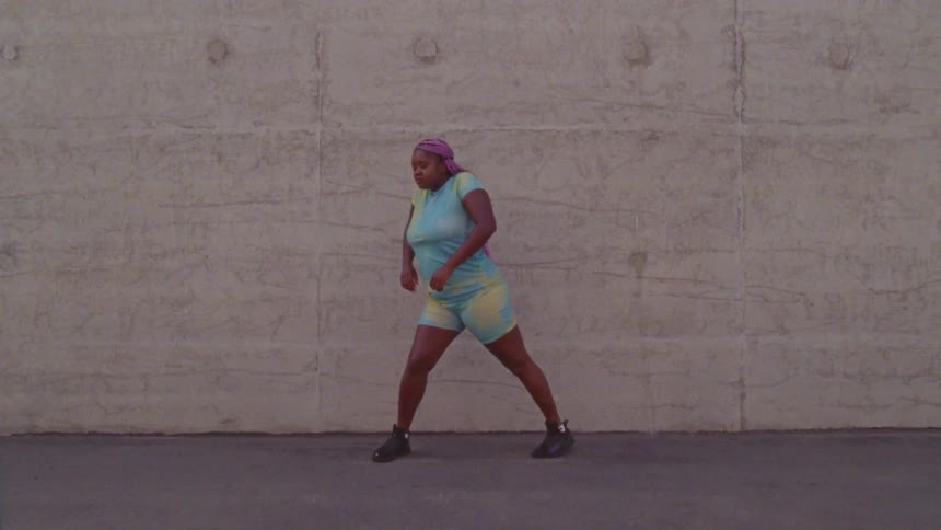 A woman in a purple head scarf and tie dye shorts and shirt does a modern dance in front of a concrete wall