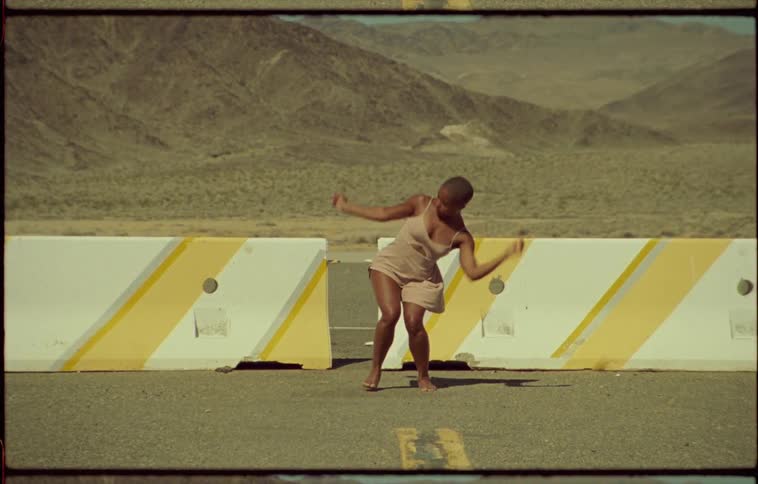 A woman dancing in a parking lot in front of dividers out in a desert like area with mountains at daytime