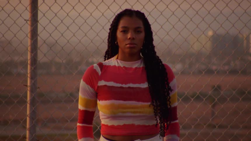 Young long haired woman standing in front of a wire fence looking at the camera at sunset