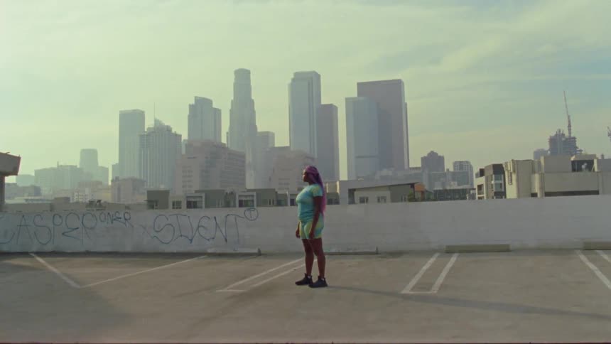 Woman stands on the top of a rooftop parking lot in a city and stares off into the distance