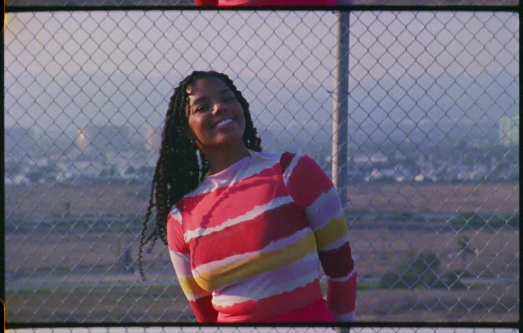 A young woman standing in front of a chain link fence makes a gesture with her body and smiles at the camera