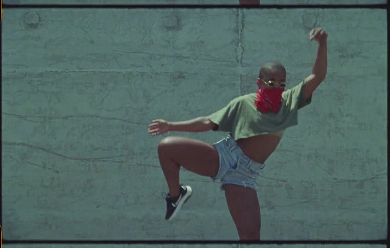 A woman in shorts with a red neck scarf dances outside in front of a concrete wall