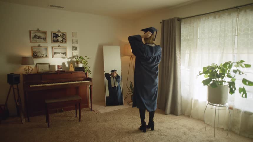 A teenage girl picks up her graduation cap and puts it on and looks in the mirror