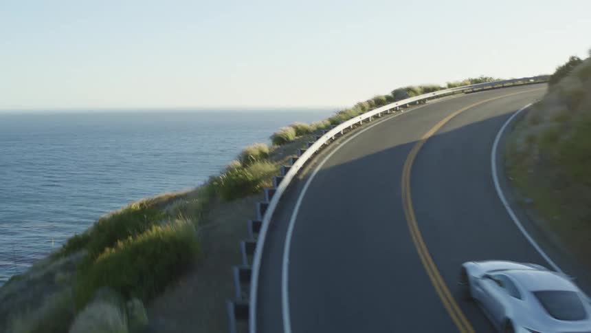Car drives along curve of highway overlooking drop off to ocean rounding hill and leaving shadow to sunshine