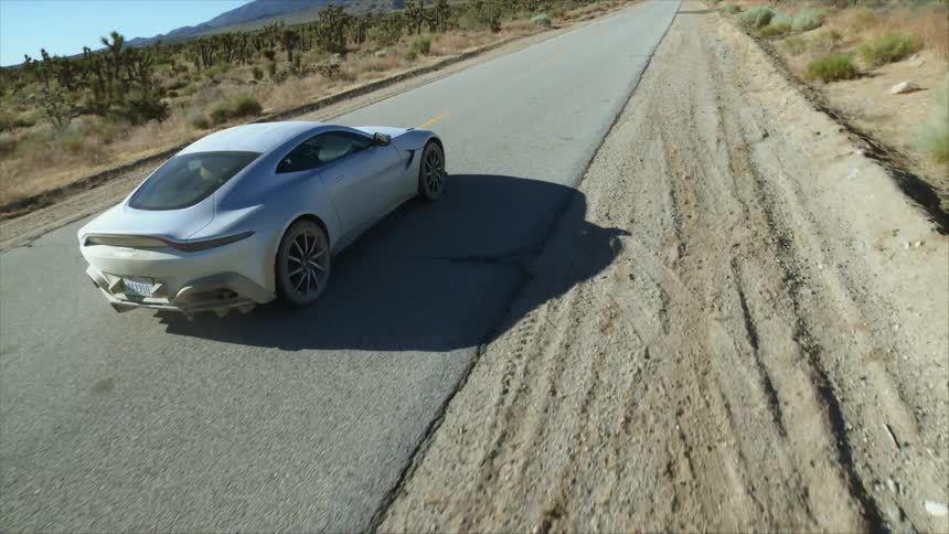 A silver car driving down a paved road in between a desert towards a mountain range at daytime