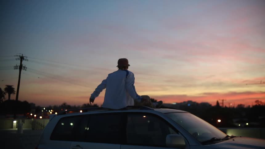 Person sitting on car roof looking toward city lights under red tinted sky waves middle fingers